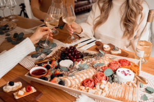 Beautiful and colorful charcuterie board of meats, cheeses, salami in the shape of roses, grapes, dips on a table setting with three women cheersing their wine glasses.